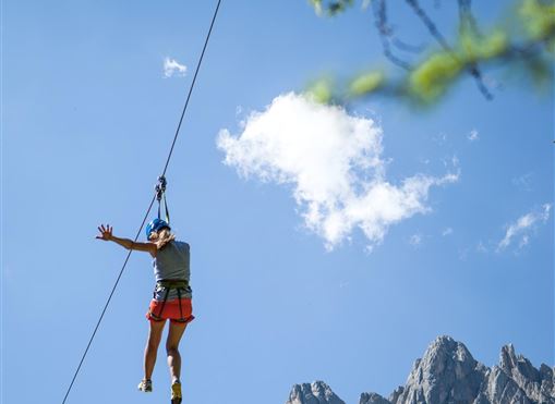 Ein Kind rutscht an einer Zipline über eine felsige Berglandschaft. Der Himmel ist blau mit wenigen Wolken.