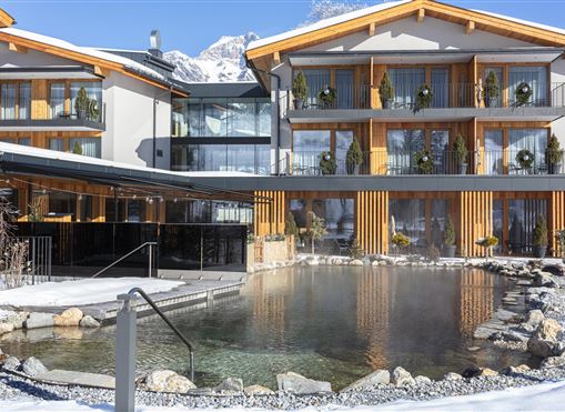 A modern hotel building with wooden cladding and large windows. In the foreground, there is a warm outdoor pool surrounded by snow-covered landscape.