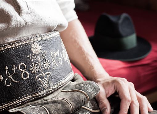 Ein traditioneller Gürtel mit aufwendiger Stickerei und eine Hand, die ihn hält. Im Hintergrund ist ein schwarzer Hut auf einem roten Bett sichtbar.