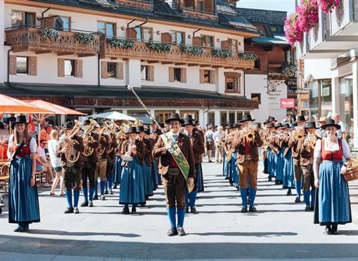 Eine Blaskapelle in traditioneller Tracht spielt in einer malerischen Straße. Im Hintergrund sind bunte Blumen und gemütliche Restaurants zu sehen.