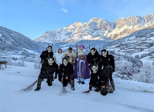 Eine Gruppe von Menschen in winterlichen Kostümen steht vor einer schneebedeckten Berglandschaft. Im Mittelpunkt steht eine Person in lila Gewand, umgeben von Figuren mit Masken und furchterregenden Kostümen.