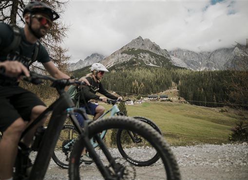 Zwei Radfahrer fahren auf einem Kiesweg in den Bergen. Im Hintergrund sind grüne Wiesen und majestätische Berge unter einem teilweise bewölkten Himmel zu sehen.