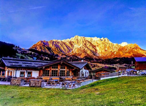 Eine malerische Berglandschaft mit schneebedeckten Gipfeln und klar blauem Himmel. Im Vordergrund befindet sich ein gemütliches Chalet mit Sitzgelegenheiten im Freien.