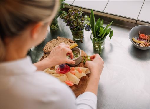 A person is preparing an appealing platter with cheese, bread, and fresh ingredients. In the background, herbs and a bowl with additional ingredients can be seen.