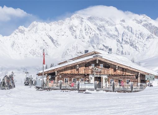 A cozy cabin surrounded by snow-covered mountains. The sky is clear and the landscape is wintry and peaceful.