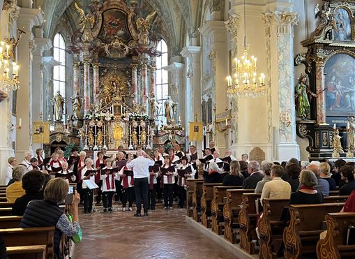 Ein Chorkonzert in einer schönen Kirche mit vielen Zuschauern. Der Chor steht in einer harmonischen Szene vor einem kunstvoll gestalteten Altar.