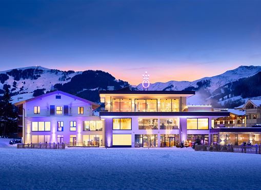 Ein modernes Gebäude in verschneiter Umgebung mit Bergblick. Die Fassade ist beleuchtet und erzeugt eine gemütliche Atmosphäre bei Dämmerung.
