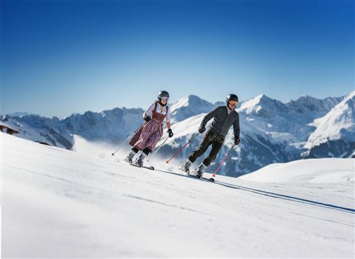 Zwei Skifahrer fahren über eine verschneite Piste in den Bergen. Der Himmel ist klar und blau, die Landschaft ist atemberaubend.