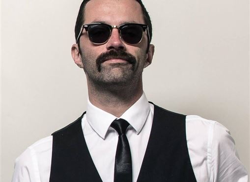 Ein stilvoll gekleideter Mann mit Sonnenbrille und Bart. Er trägt eine Weste und eine Krawatte vor einem neutralen Hintergrund.