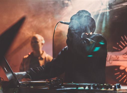 Ein DJ steht an den Turntables und spielt Musik, während er ein Mikrofon hält. Im Hintergrund ist eine zweite Person zu sehen.