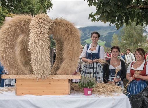 Drei Frauen in traditioneller Tracht stehen vor einem kunstvoll dekorierten Strohkranz. Im Hintergrund sind grüne Landschaften und ein bewölkter Himmel zu sehen.