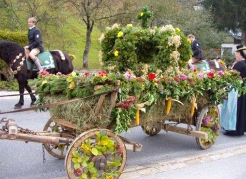 Ein mit Blumen und Gras geschmückter Wagen fährt entlang einer Straße. Im Hintergrund sieht man Reiter auf Pferden.
