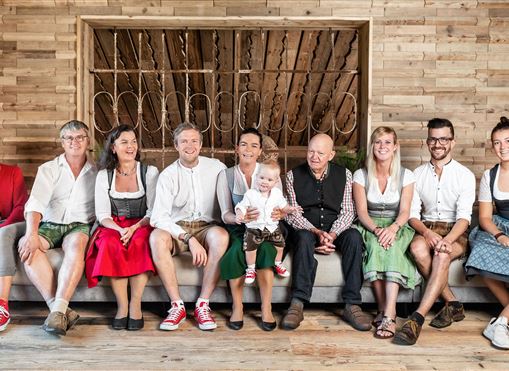 Eine Gruppe von Menschen sitzt auf einem Sofa in traditioneller Tracht. Sie lächeln und wirken freundlich, in einem rustikalen Holzinterieur.