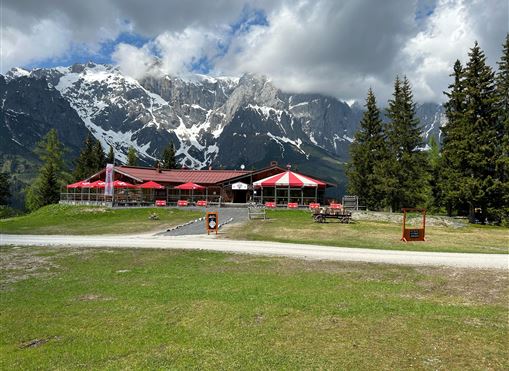 Eine Berghütte mit rotem Dach und Sonnenschirmen steht vor einer beeindruckenden Bergkulisse. Umgeben von grünen Wiesen und hohen Bäumen lädt sie zur Entspannung ein.
