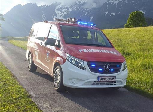 Ein Feuerwehrfahrzeug steht auf einer Straße in einer malerischen Landschaft. Im Hintergrund sind Berge und ein bewölkter Himmel sichtbar.