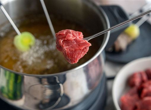 Ein Stück rotes Fleisch wird mit einer Gabel in einen Topf mit heißem Öl getaucht. Im Hintergrund sind weitere Fleischstücke auf einem Teller zu sehen.
