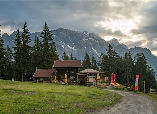 Ein malerisches Berghaus umgeben von grünen Wiesen und hohen Bäumen. Im Hintergrund sind majestätische Berge mit schneebedeckten Gipfeln und einem bewölkten Himmel zu sehen.
