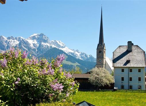 Eine malerische Landschaft mit schneebedeckten Bergen und einem klaren blauen Himmel. Im Vordergrund steht ein schönes Dorfhaus mit einer Kirche und blühenden Pflanzen.