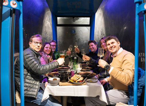Eine Gruppe von Menschen sitzt an einem Tisch und hebt Gläser zum Anstoßen. Das Ambiente ist gemütlich mit einer blauen Beleuchtung.
