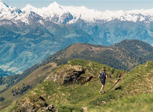Ein Läufer erkundet eine malerische Berglandschaft. Im Hintergrund sind schneebedeckte Gipfel und grüne Täler zu sehen.