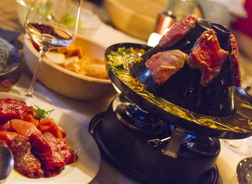 Ein geschmackvolles Fondue-Gericht mit Fleisch und Beilagen auf einem Tisch. Im Hintergrund ist ein Glas Wein zu sehen, was eine gemütliche Atmosphäre schafft.