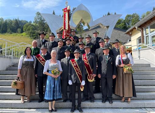 A group of people in traditional attire stands on the steps in front of modern architecture. They are smiling and holding flags as well as flowers in their hands.