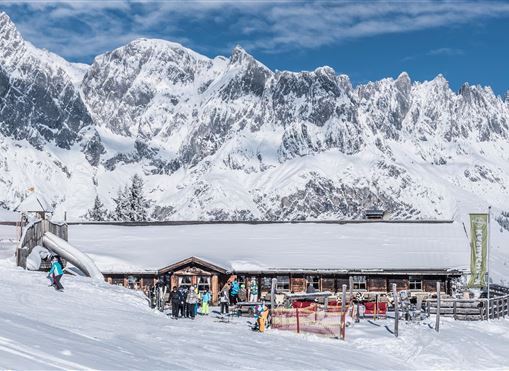Eine Skihütte in den Alpen, umgeben von schneebedeckten Bergen. Skifahrer genießen den sonnigen Tag im Schnee.