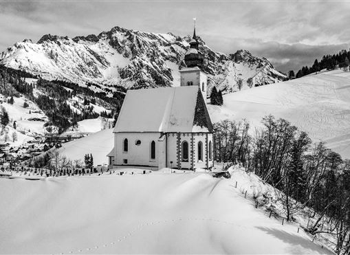 Eine charmante Kirche steht inmitten einer verschneiten Landschaft mit Bergen im Hintergrund. Die Szenerie strahlt Ruhe und Frieden aus.
