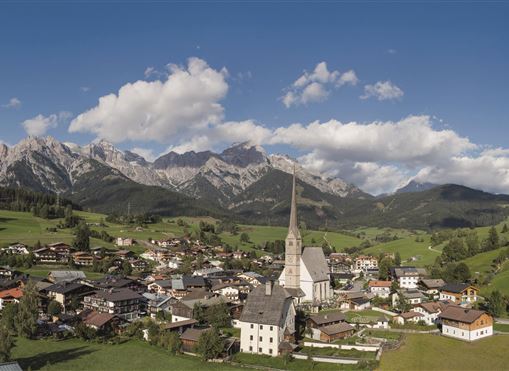 Ein malerisches Dorf mit einer Kirche und traditionellen Häusern. Im Hintergrund erheben sich majestätische Berge unter einem klaren blauen Himmel.