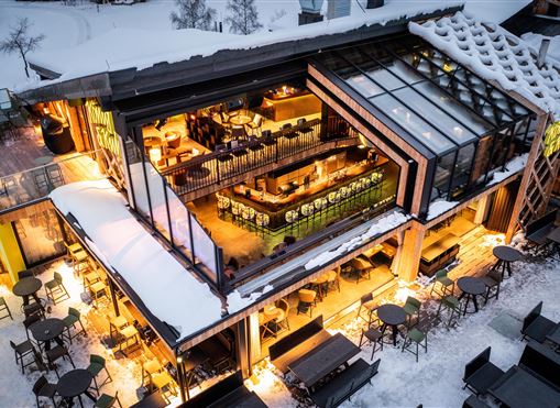 Ein modernes Restaurant mit großen Fenstern und warmem Licht in einem schneebedeckten Umfeld. Gemütliche Sitzbereiche im Freien laden zum Verweilen ein.