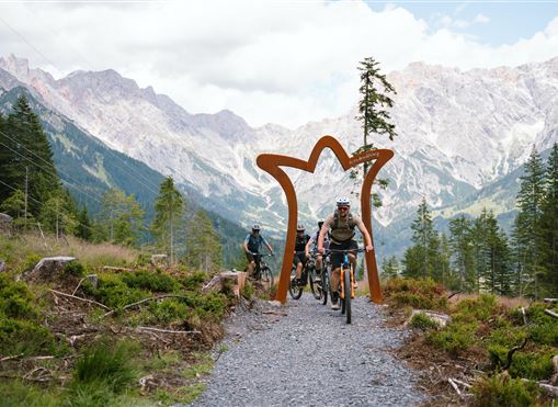 Eine Gruppe von Radfahrern fährt durch einen Naturpfad in den Bergen. Im Hintergrund sind beeindruckende Berglandschaften und ein kunstvolles Tor zu sehen.