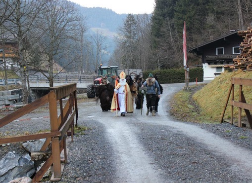 Eine Gruppe von Personen geht auf einem Schotterweg, begleitet von Pferden. Im Hintergrund sind Bäume und ein Gebäude zu sehen.