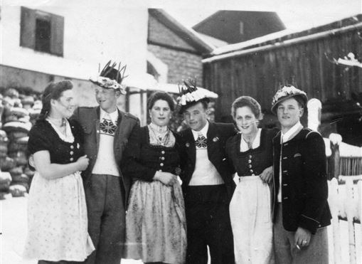 A group of six people are wearing traditional costumes and hats. They are standing smiling in front of a rural background with buildings.