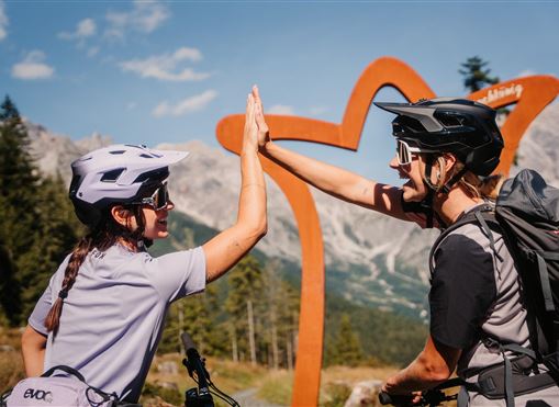 Zwei Mountainbiker fahren durch einen Waldweg mit einer kunstvollen Metallskulptur im Vordergrund. Im Hintergrund sind die beeindruckenden Berge und eine bewaldete Landschaft zu sehen.