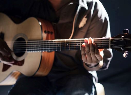 A musician is playing guitar in the dark background. The musician's hand is strumming the strings of the acoustic guitar.
