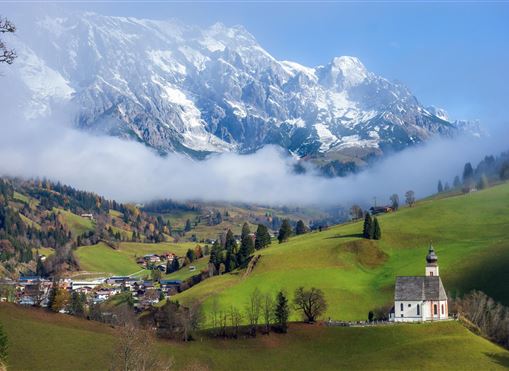 Eine malerische Landschaft mit grünen Wiesen und schneebedeckten Bergen im Hintergrund. Im Vordergrund steht eine kleine Kirche in der Nähe eines Dorfes.
