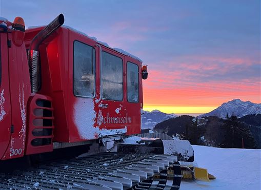 Eine rote Pistenraupe steht im Schnee bei Sonnenuntergang. Im Hintergrund sind Berge und ein farbenfroher Himmel zu sehen.