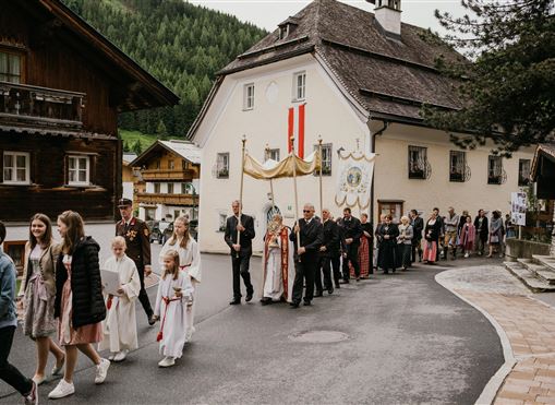 Eine Feierlichkeit mit einer Prozession, bei der Menschen in festlicher Kleidung entlang einer Straße gehen. Im Hintergrund sind traditionelle Gebäude und eine grüne Landschaft zu sehen.