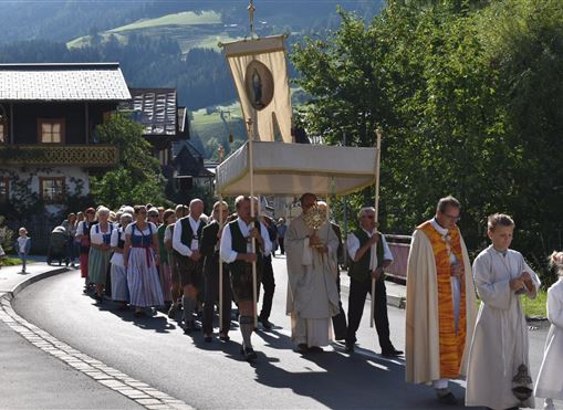 Eine festliche Prozession mit Menschen in traditioneller Kleidung, die durch eine malerische Straße gehen. Im Vordergrund trägt eine Person eine großformatige Monstranz.