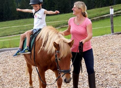 Ein Kind reitet fröhlich auf einem Pony und hat die Arme ausgebreitet. Eine Frau begleitet das Pony und schaut lächelnd auf das Kind.