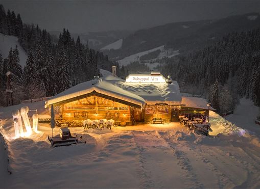 Eine gemütliche Berghütte im Schnee bei Nacht. Warmes Licht strahlt aus dem Gebäude, umgeben von verschneiten Bäumen und Bergen.