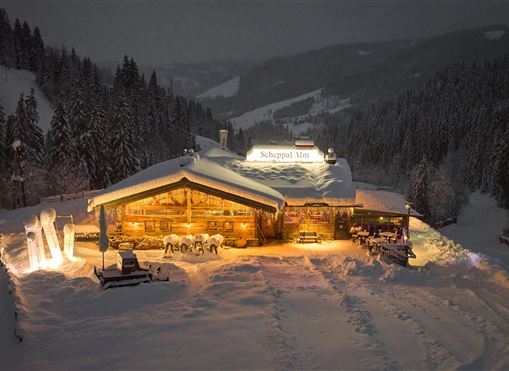 Eine chaletartige Hütte ist von Schnee umgeben und sanft beleuchtet. Die winterliche Landschaft und die Berge im Hintergrund schaffen eine gemütliche Atmosphäre.