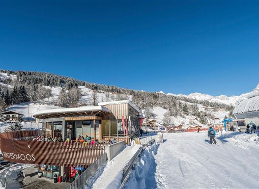 A cozy mountain cabin in a snowy winter scene. In the background, snow-covered mountains and a clear blue sky can be seen.