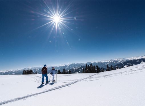 Zwei Skifahrer stehen auf einer schneebedeckten Fläche unter einem strahlend blauen Himmel. Die Sonne scheint sehr hell und die Berge sind im Hintergrund sichtbar.