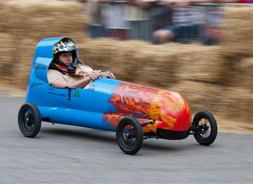 Ein Rennfahrer sitzt in einem blauen Seifenkistenauto mit Flammendekor. Das Fahrzeug fährt schnell auf einer Strecke, umgeben von Strohballen und Zuschauern.