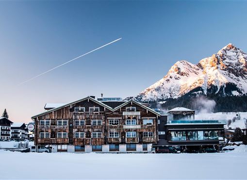 Ein charmantes Holzhaus inmitten einer schneebedeckten Landschaft. Im Hintergrund erheben sich majestätische Berge under einem klaren Himmel.
