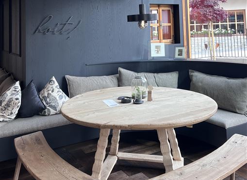 A cozy seating area with a round wooden table and a corner bench. Large windows provide plenty of light and a view of the outside world.