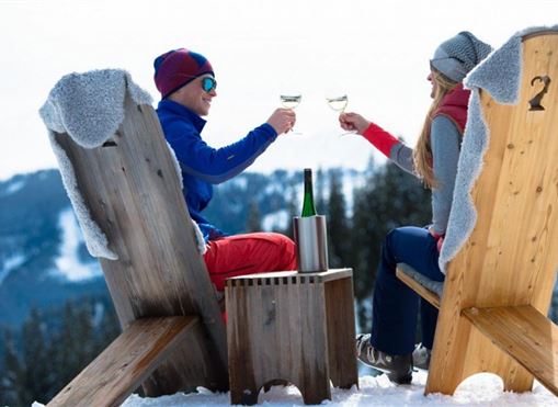 Ein Paar sitzt auf Holzstühlen im Schnee und prostet sich mit Gläsern zu. Im Hintergrund sind schneebedeckte Berge und ein klarer Himmel zu sehen.
