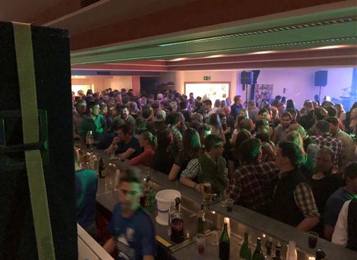 A lively party with many people standing at the bar and celebrating. The surroundings are brightly lit, and there is a cheerful atmosphere.