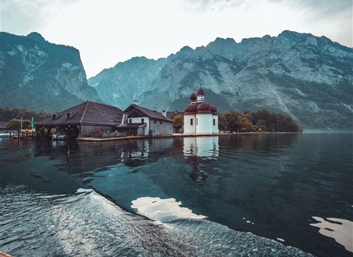 Eine ruhige Seenlandschaft mit einem kleinen weißen Gebäude am Wasser. Im Hintergrund erheben sich majestätische Berge.